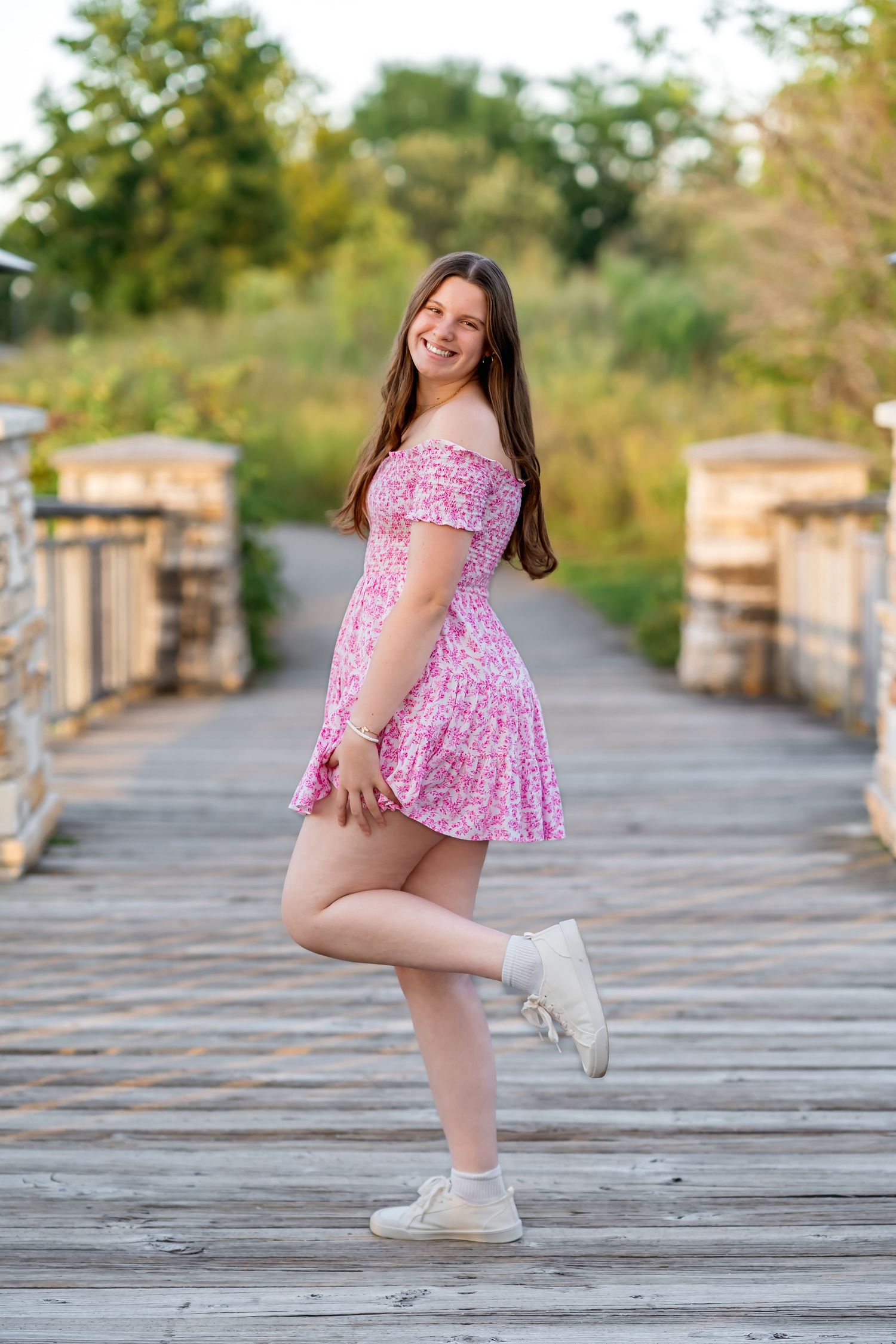 A person in a pink floral dress and white sneakers poses playfully on a wooden boardwalk surrounded by stone pillars.