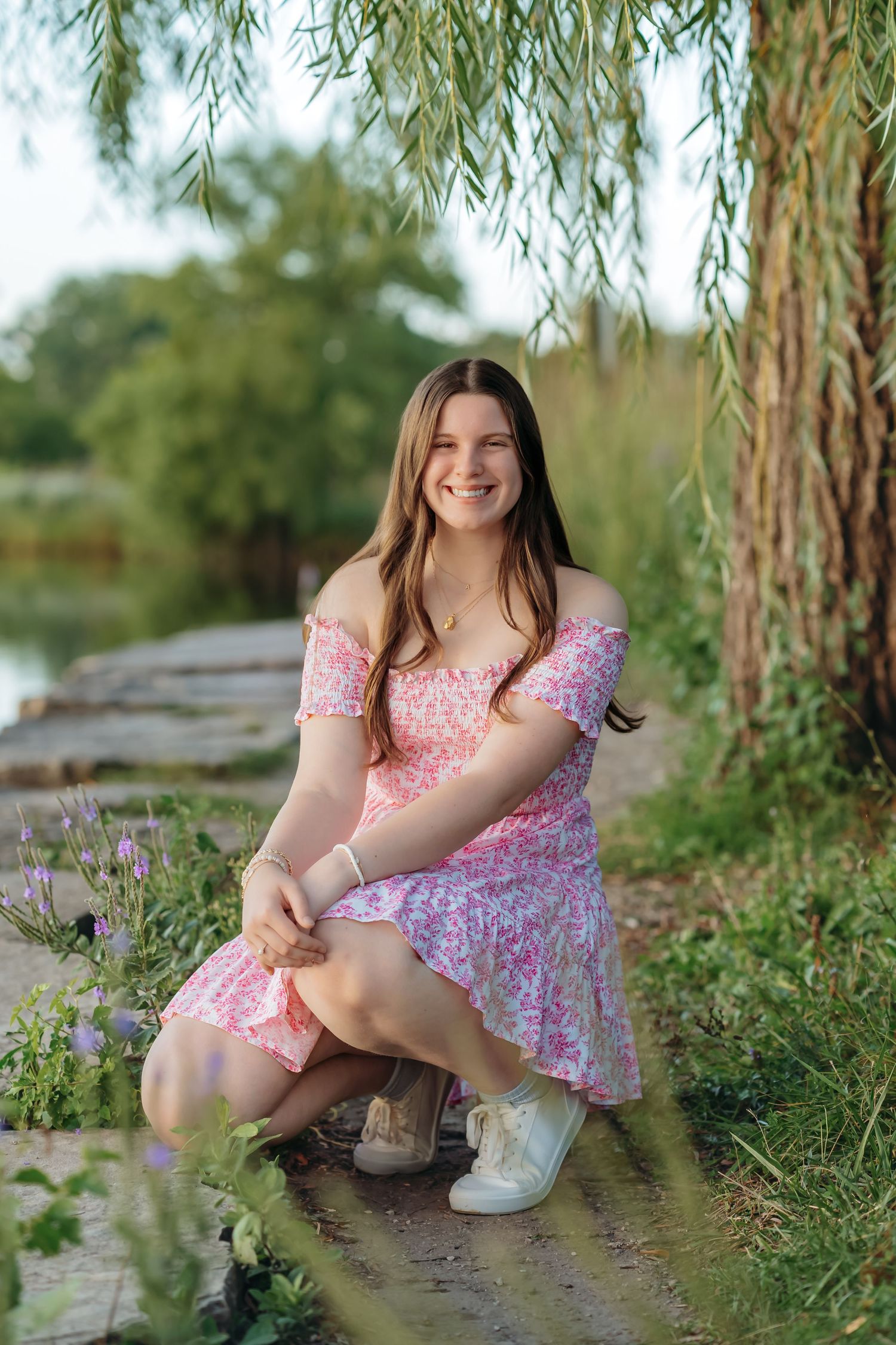 A person in a pink floral dress poses under a weeping willow tree next to blooming purple wildflowers.