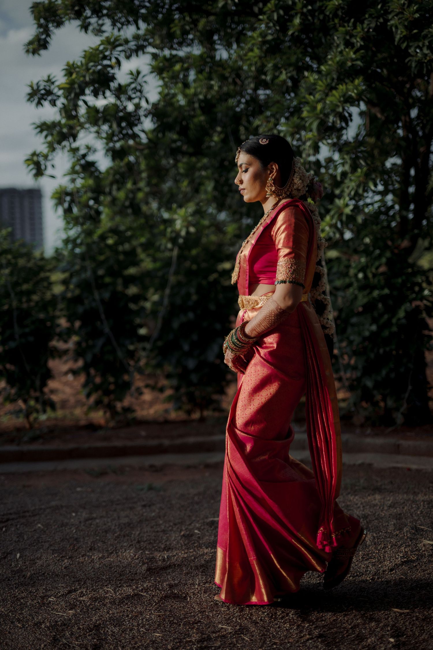 Traditional bridal portrait in a red silk saree against a natural outdoor setting.
