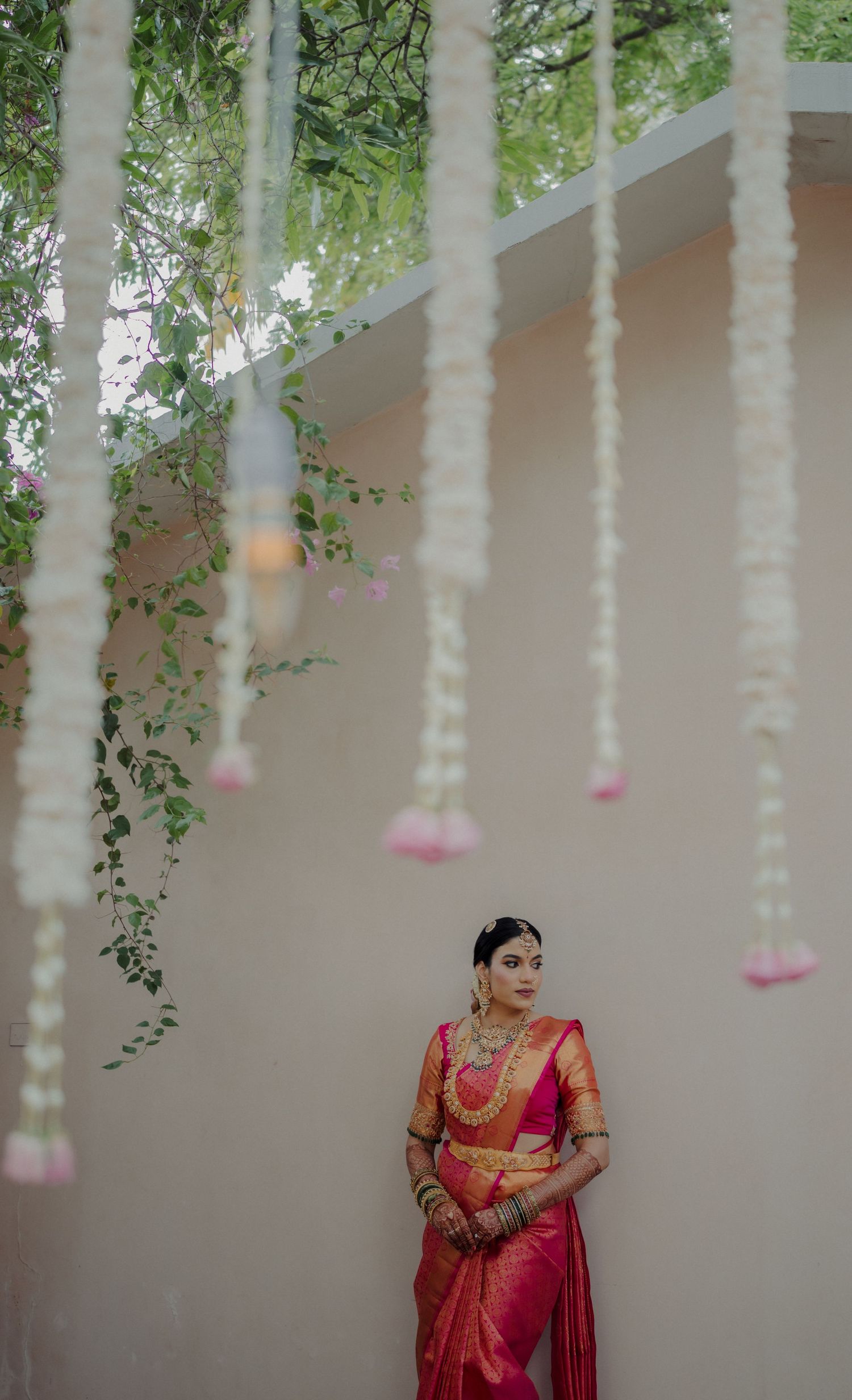 Traditional South Indian bride in red silk saree poses elegantly under hanging floral decorations and strings.