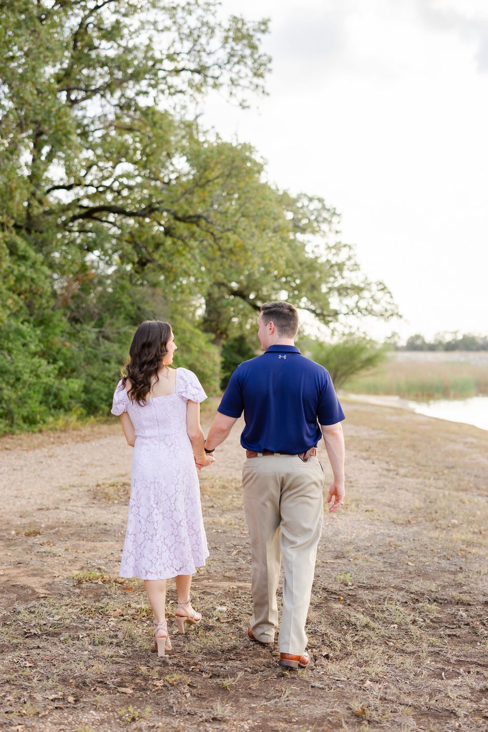 A couple walks hand in hand down a dirt path beside trees and grassy fields on a bright sunny day.