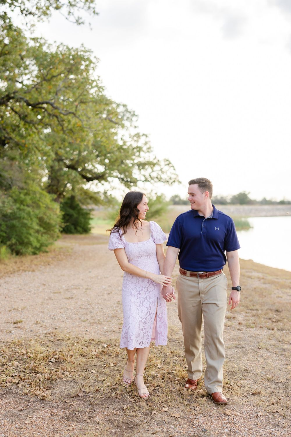 A couple in casual attire walks together on a dirt path surrounded by greenery on a sunny day.
