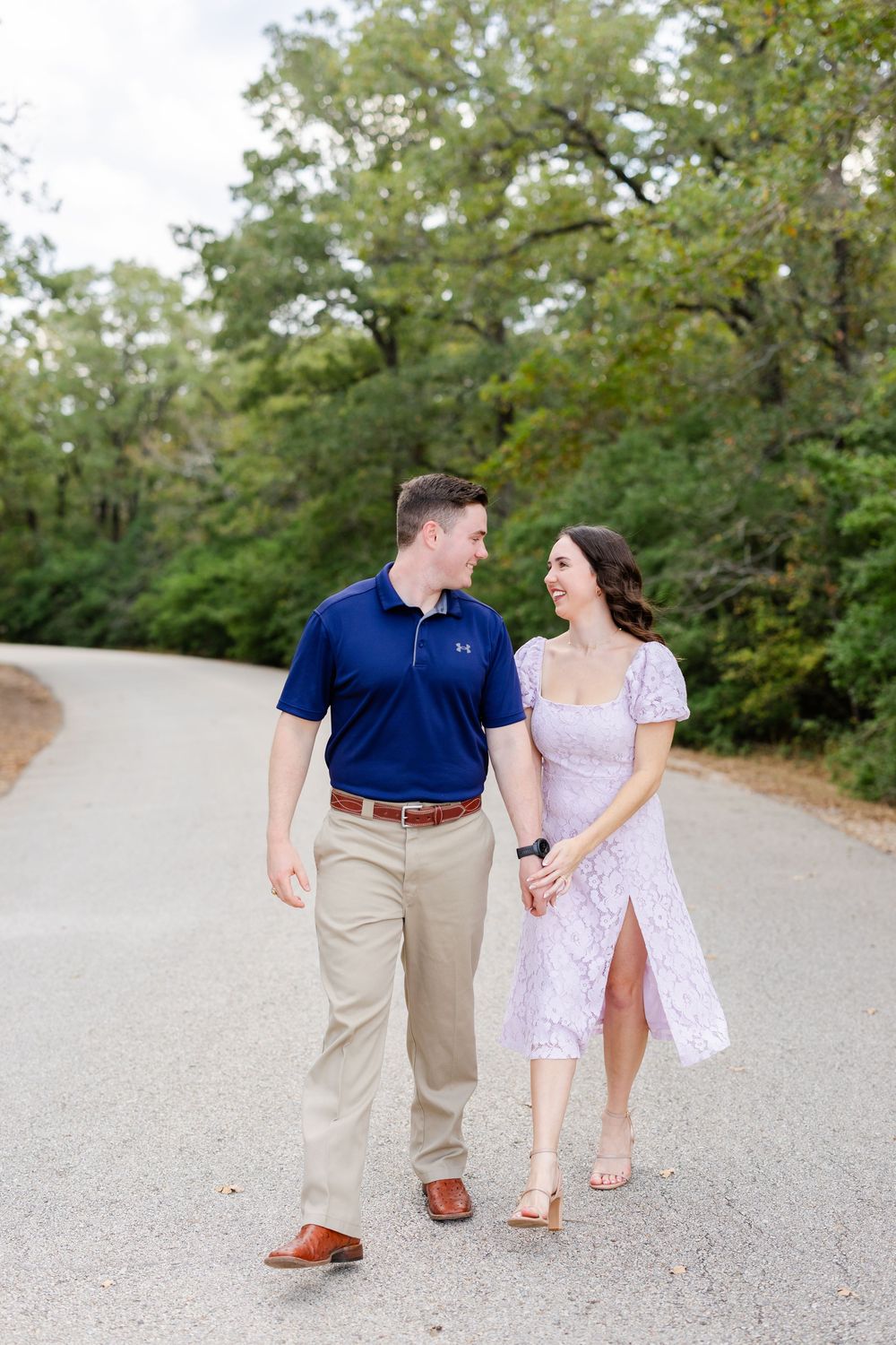 A couple in coordinated navy and white outfits walk hand in hand down a tree-lined gravel path.