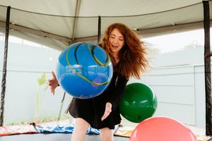A person with long red hair dances energetically with colorful balloons in a white tent-like structure.