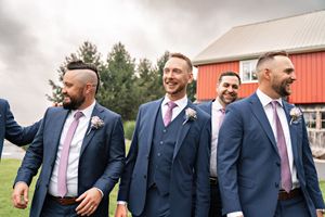 Groomsmen in navy suits with pink ties laugh together while standing near a red barn on wedding day.