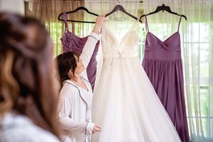 Purple bridesmaid dresses and white bridal gown hanging on display in a sunlit room with sheer curtains.