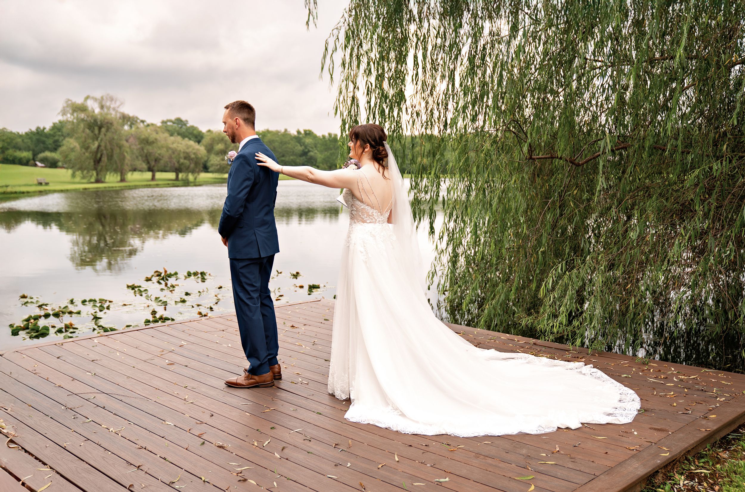 Newlyweds stand on a wooden dock surrounded by weeping willow trees at a lakeside wedding venue.