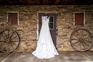 Rustic stone wall backdrop with vintage wagon wheels and an elegant white wedding dress displayed in natural light.