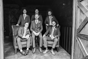 Groomsmen in navy suits sit together on rustic wooden step laughing and sharing a candid moment.