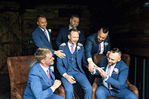 Groomsmen in blue suits gathered outdoors sharing jokes and laughter on weathered wooden steps.