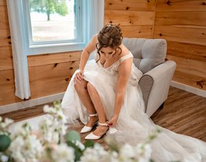 A bride in a white wedding dress adjusts her shoes while sitting in an armchair inside a rustic cabin setting.