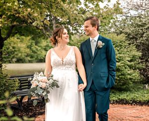 Romantic wedding couple strolls through a garden pathway with blooming flowers and trees on a sunny day.