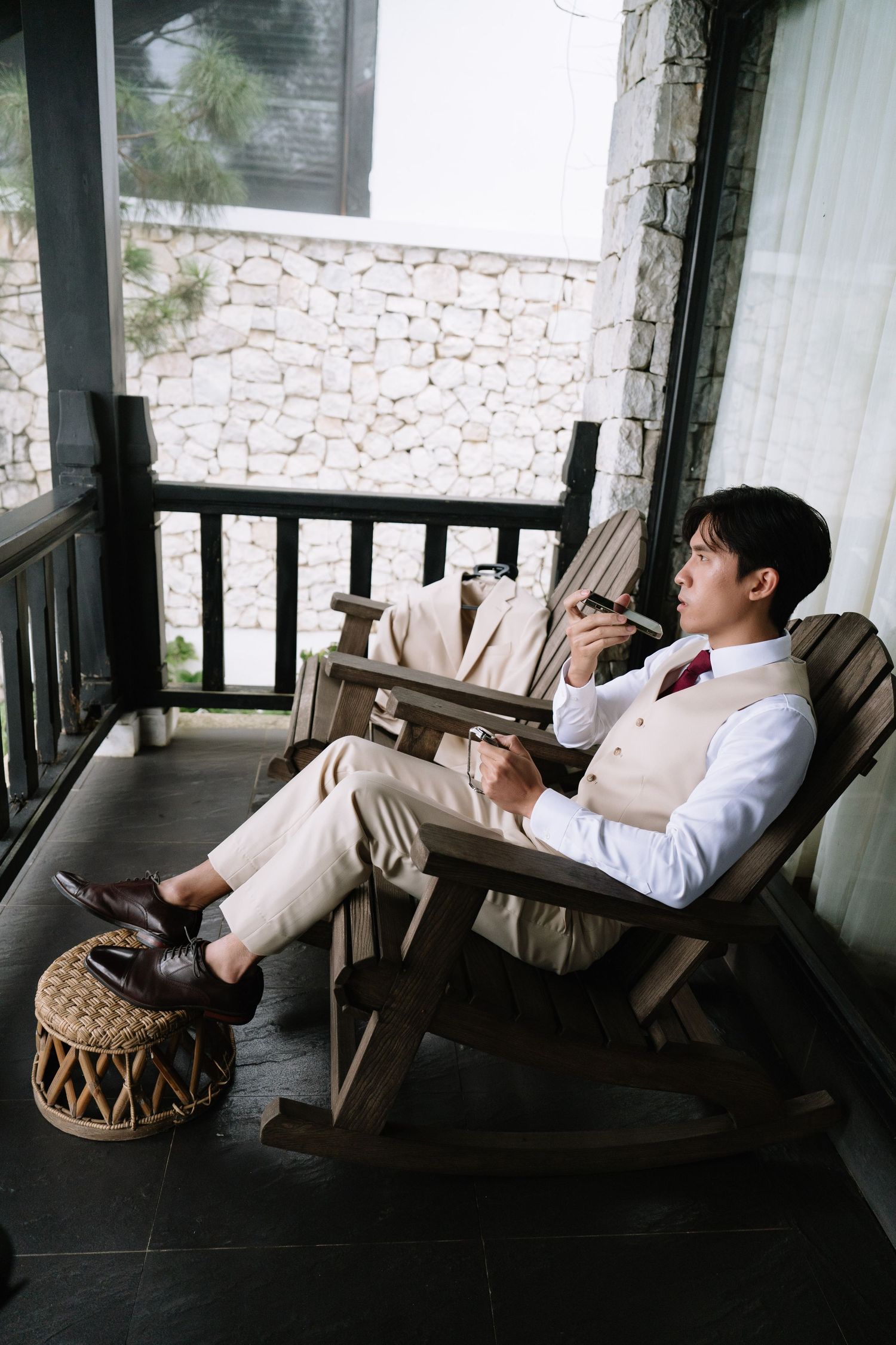 Groom in beige vest suit relaxing in wooden chair on a rustic porch holding a camera.