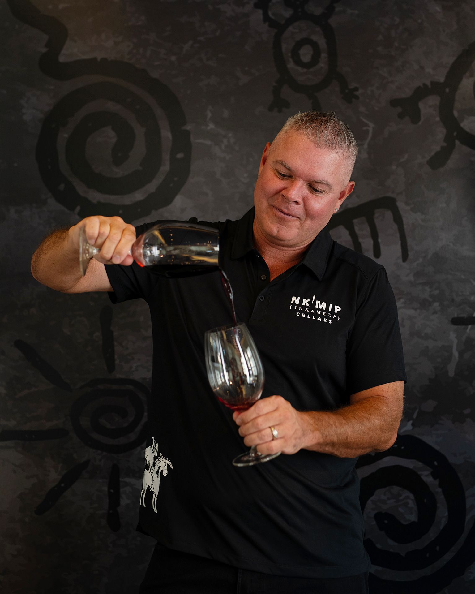 Man in black shirt pouring red wine into a glass against a textured backdrop.