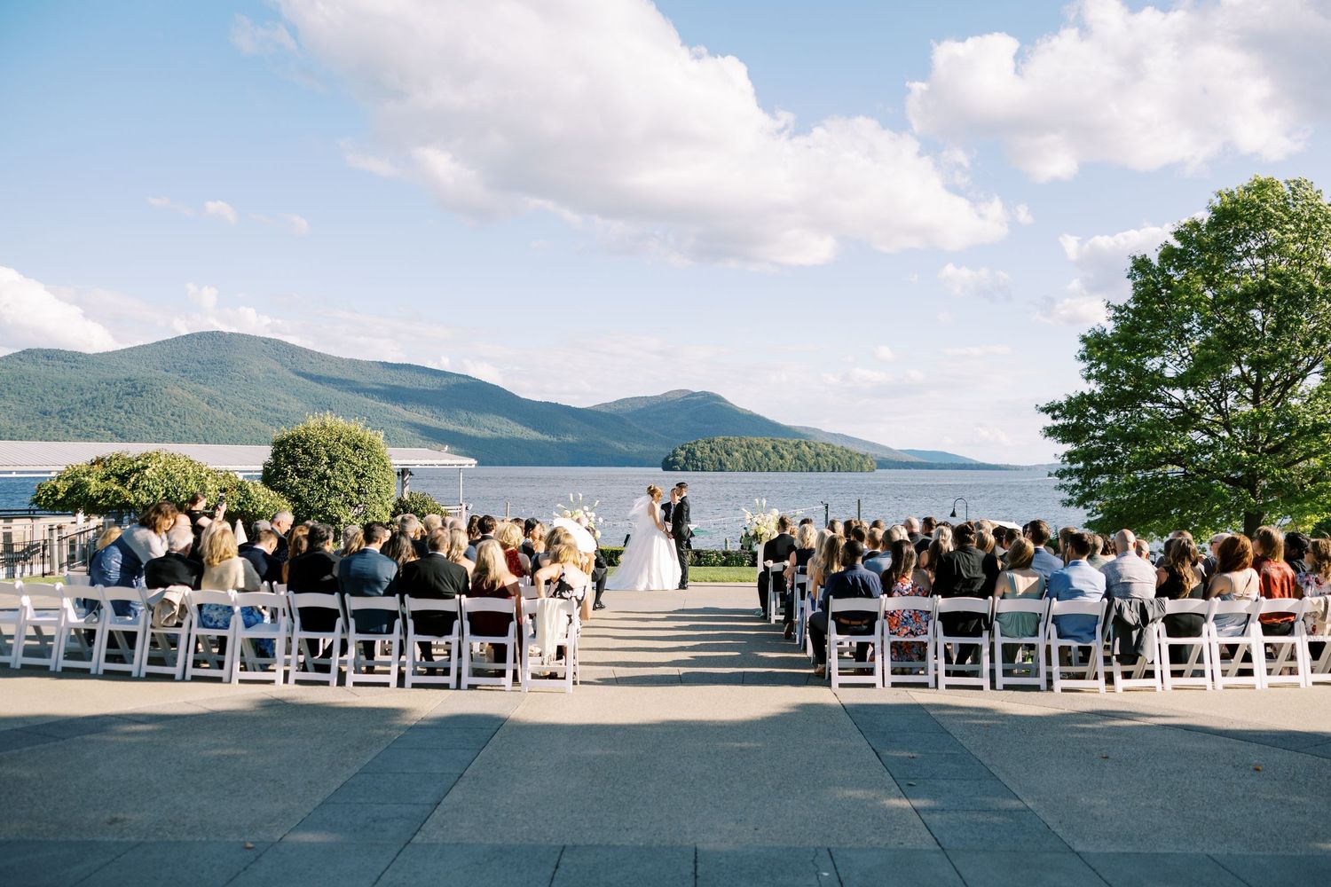 Row of white chairs arranged overlooking mountain lake with blue skies and fluffy clouds at outdoor wedding ceremony.