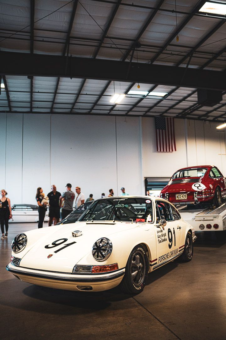 Three vintage white Porsche race cars are displayed in a row inside a dimly lit warehouse showroom.