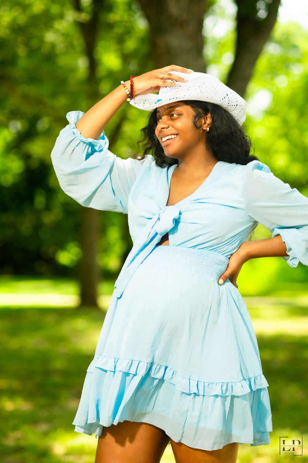 Playful poses in a light blue summer dress against a green park background with trees.