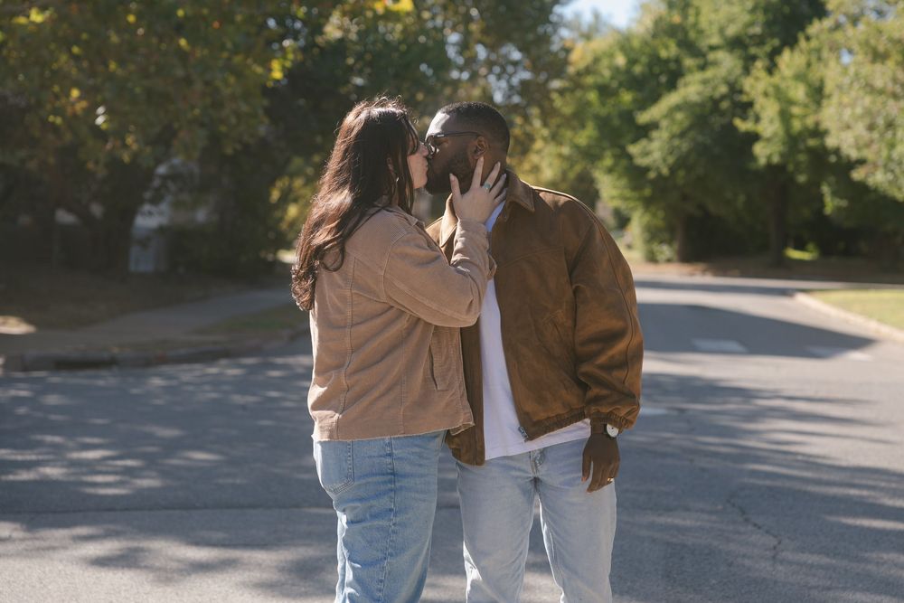 A couple in casual fall attire embraces while walking together on a tree-lined path on a sunny autumn day.