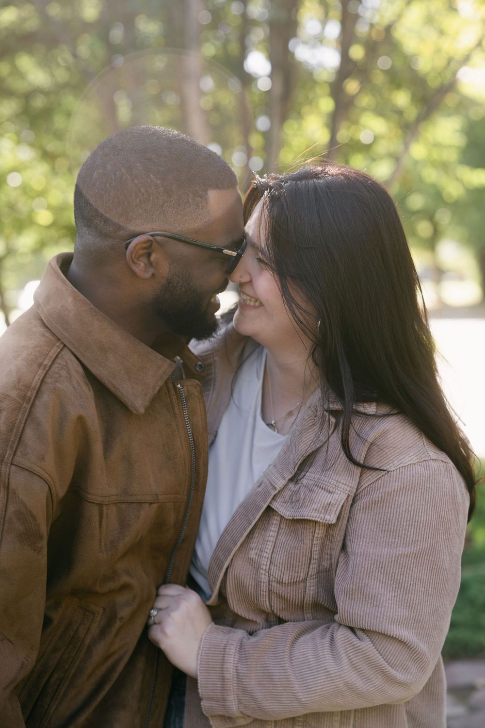 A romantic couple in brown jackets share an intimate moment outdoors with sunlit trees in the background.