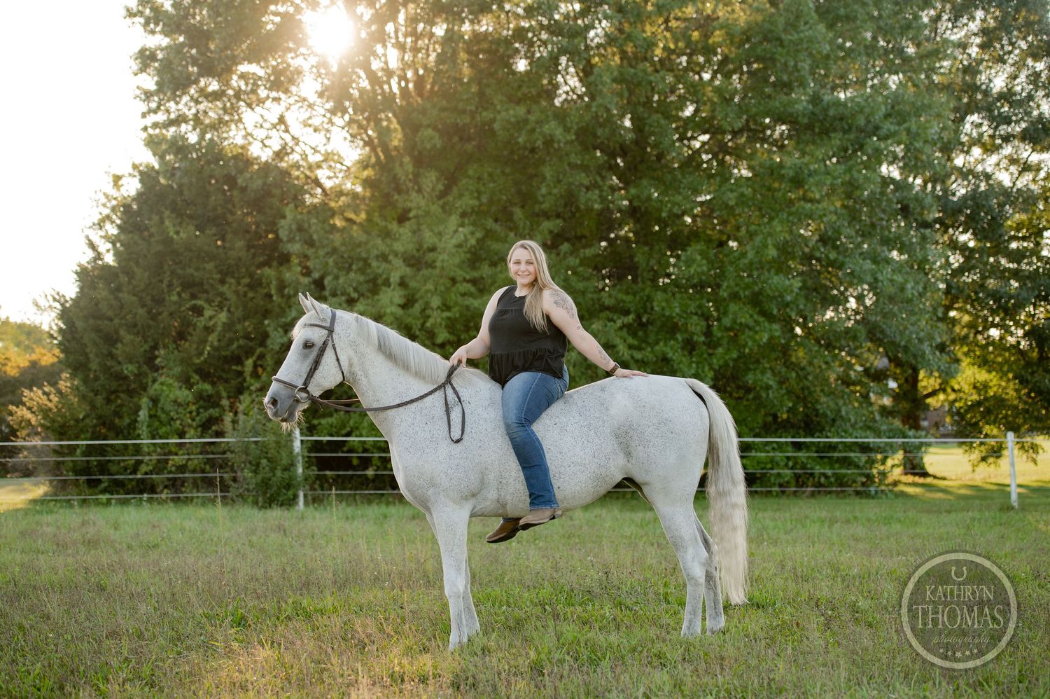 A person rides a white horse in a fenced field during golden hour.