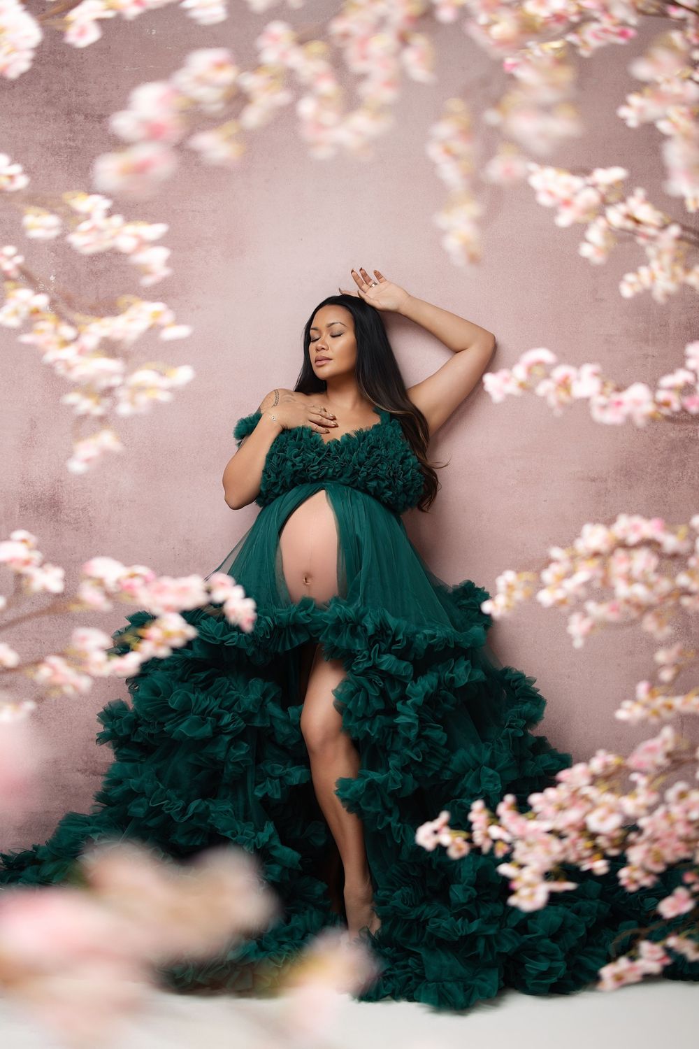 Pregnant woman in emerald green ruffled gown poses gracefully among pink cherry blossoms on a mauve background.