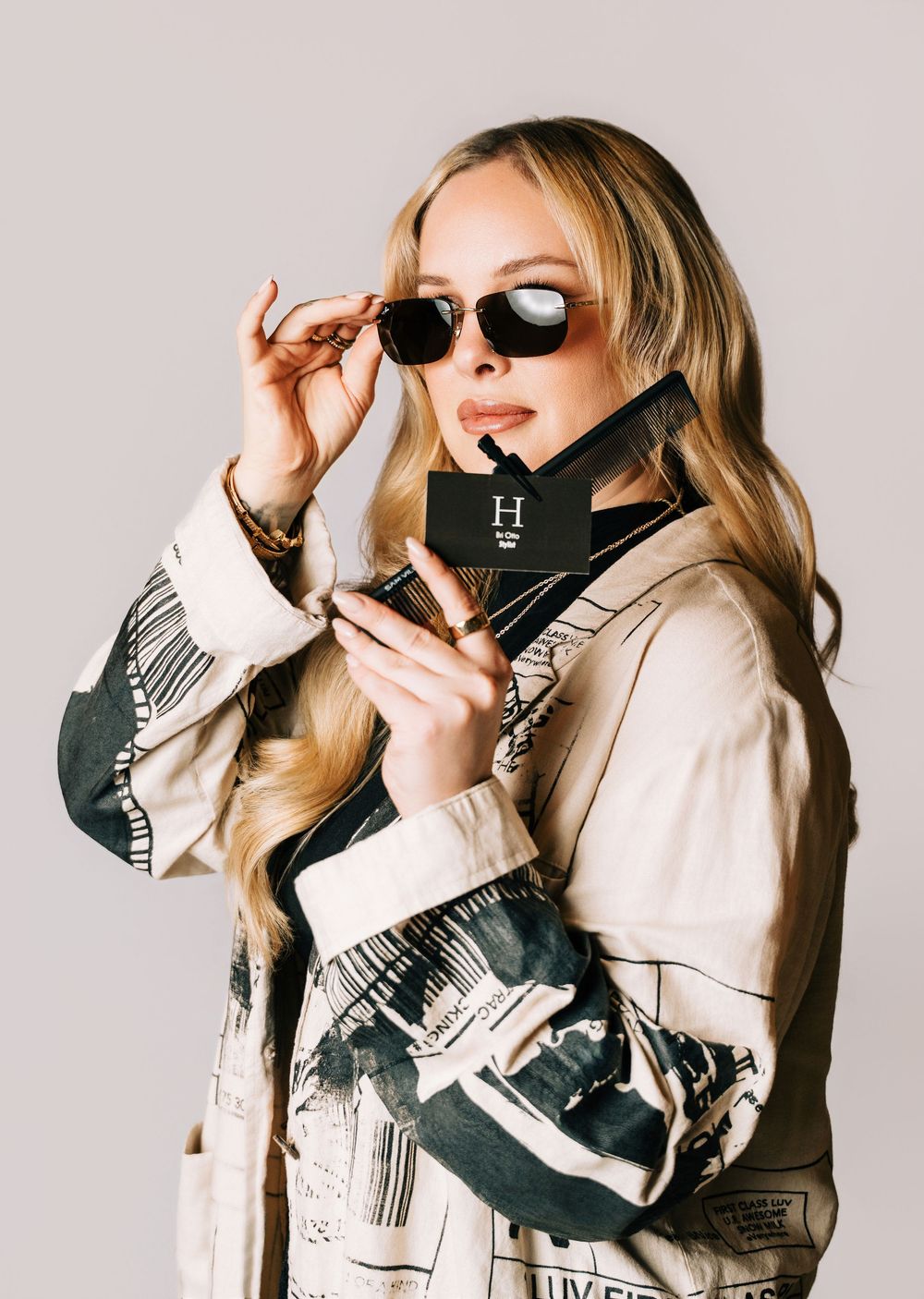 Blonde woman in printed jacket adjusts sunglasses while holding a black card in a stylish fashion portrait.