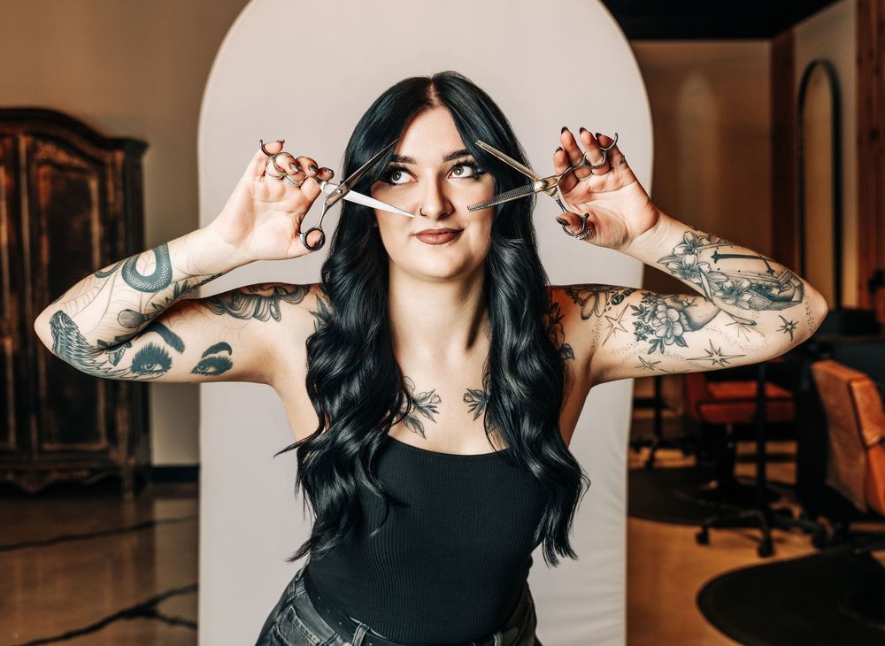 Tattooed female hairstylist holding scissors near face in modern salon, wearing black top with long dark hair.