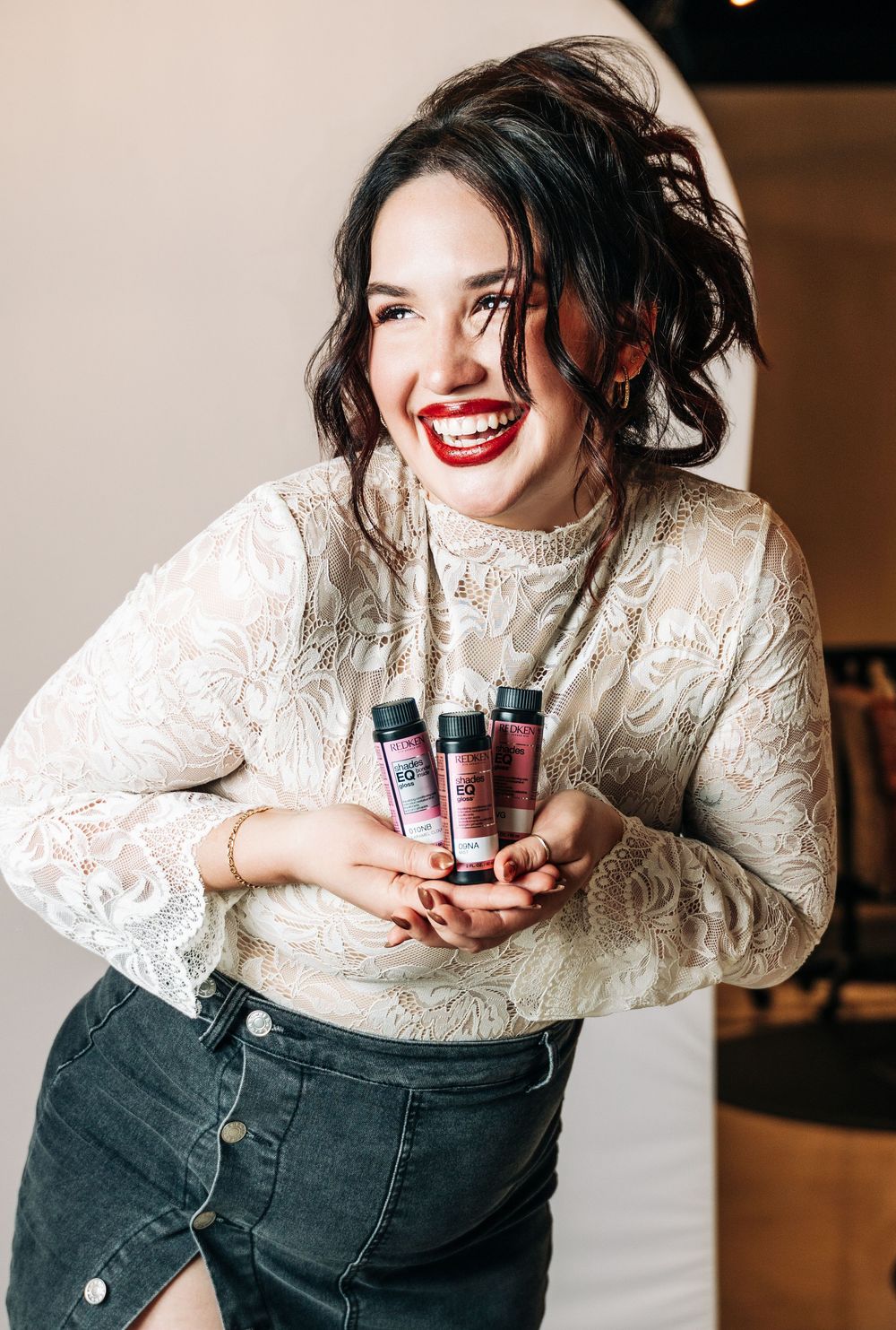 Smiling woman in white lace top holding red hair care products, wearing bold red lipstick and dark jeans.