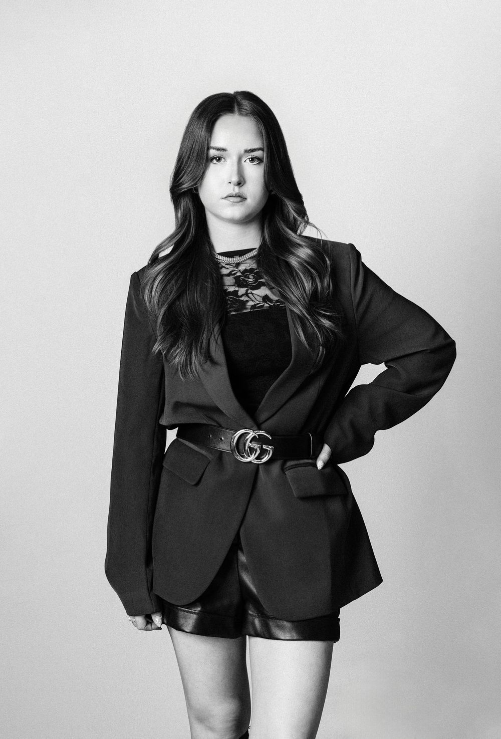 Young woman in black blazer, Gucci belt, and leather shorts poses confidently in a black and white studio photo.