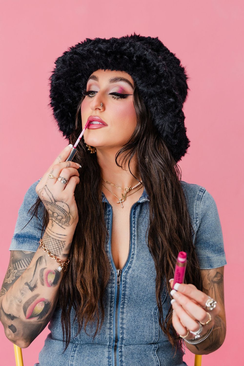 Person with long dark hair and tattoos poses against pink background while applying lipstick in sequence of photos.
