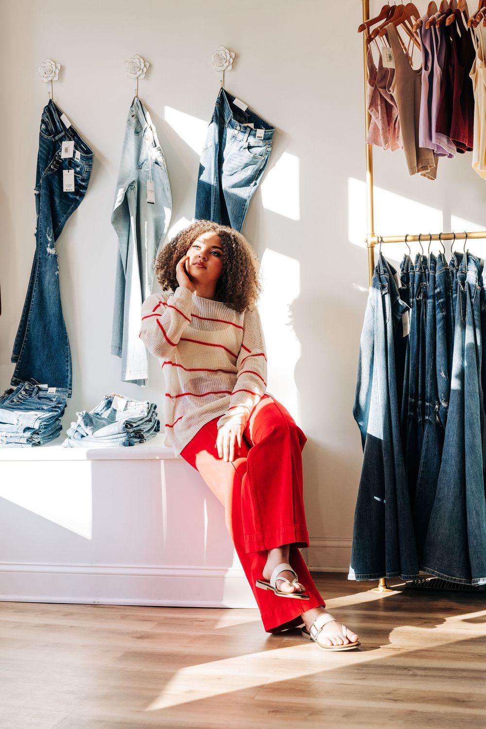 Woman in red pants and striped sweater sitting in a boutique clothing store surrounded by denim jeans.