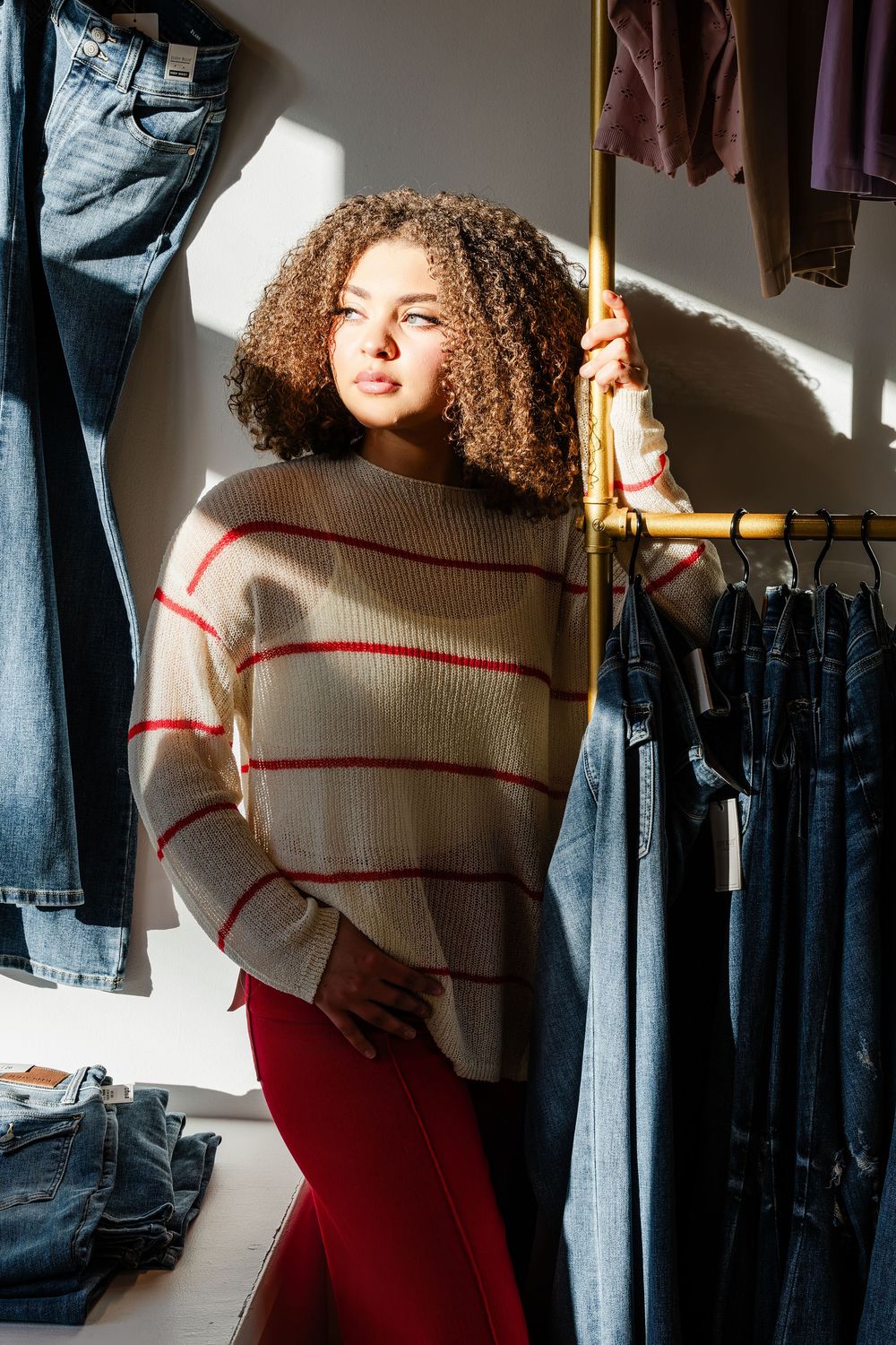 Person wearing beige sweater with red stripes and burgundy pants stands among hanging clothes in a closet.