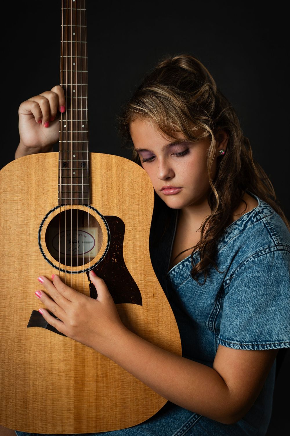 Young girl in denim vest leaning against acoustic guitar on dark background.