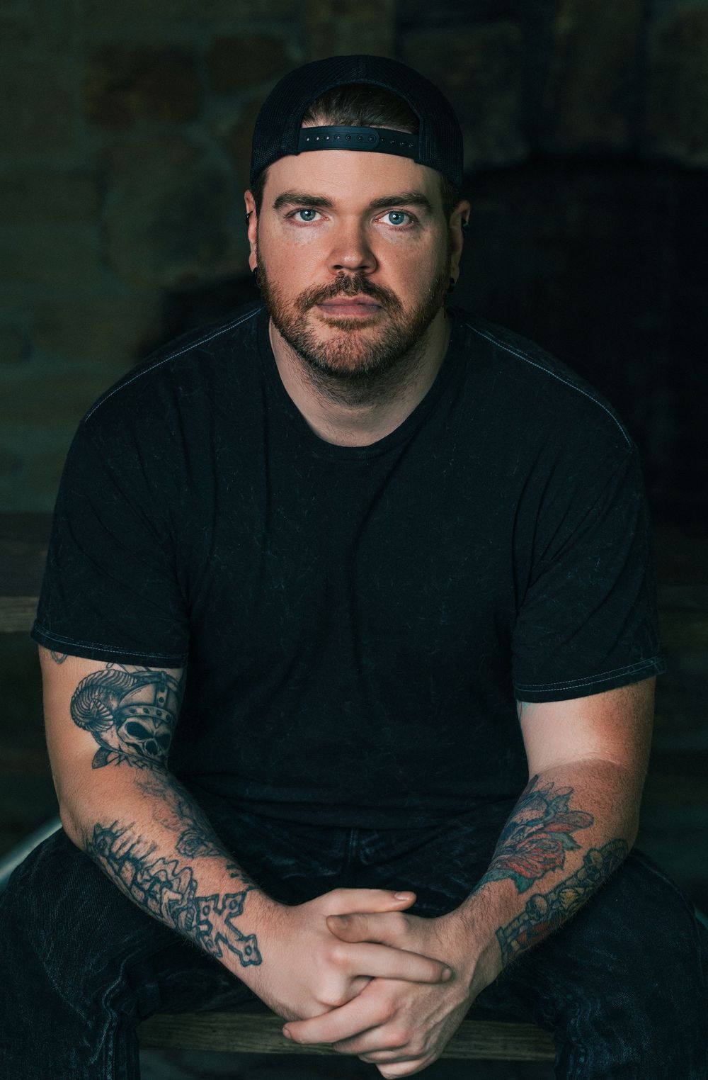 Tattooed man in black t-shirt and backwards cap sits with hands clasped, posing for a portrait photo.