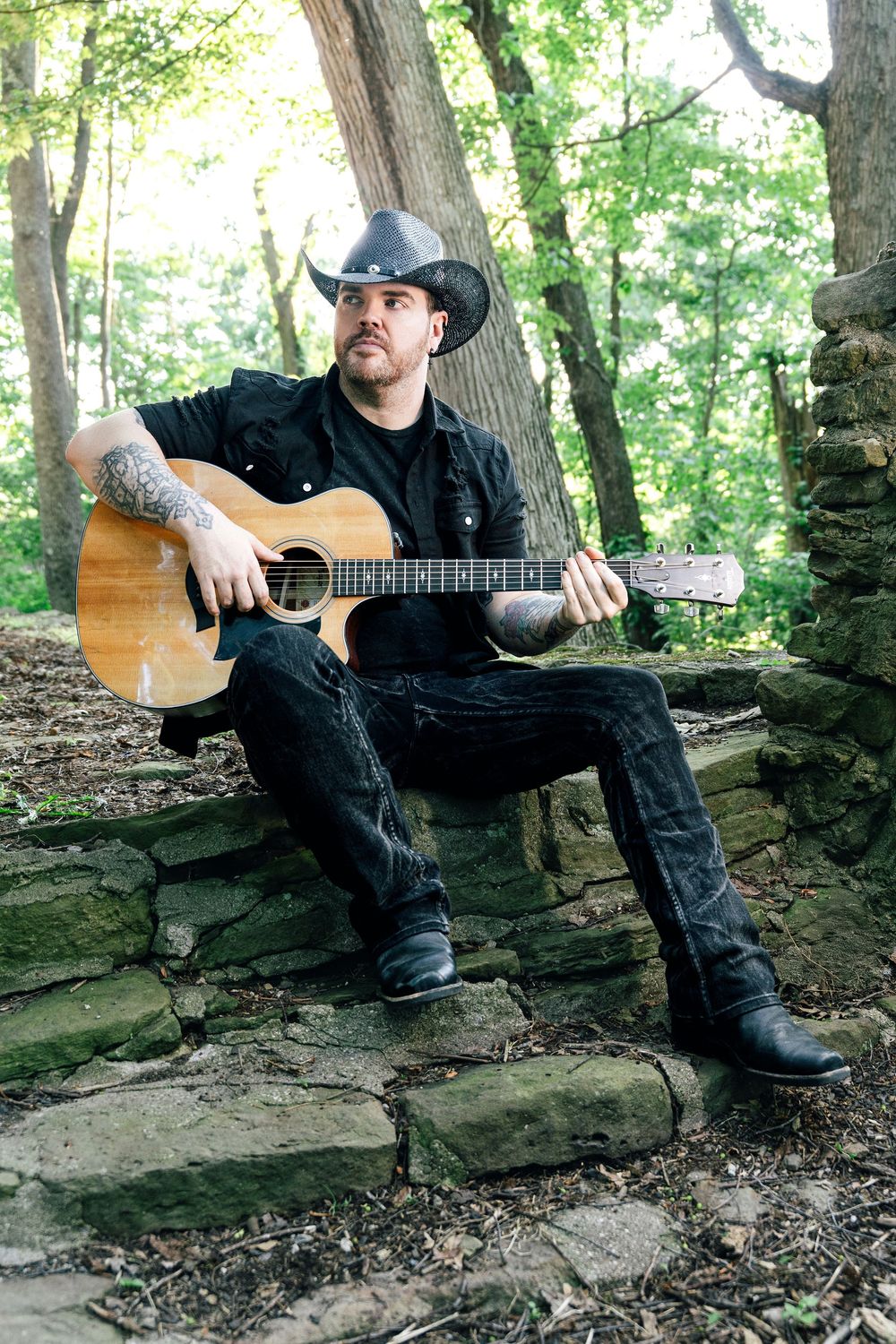 A man in a black cowboy hat and outfit sits on stone steps in a forest, playing an acoustic guitar.