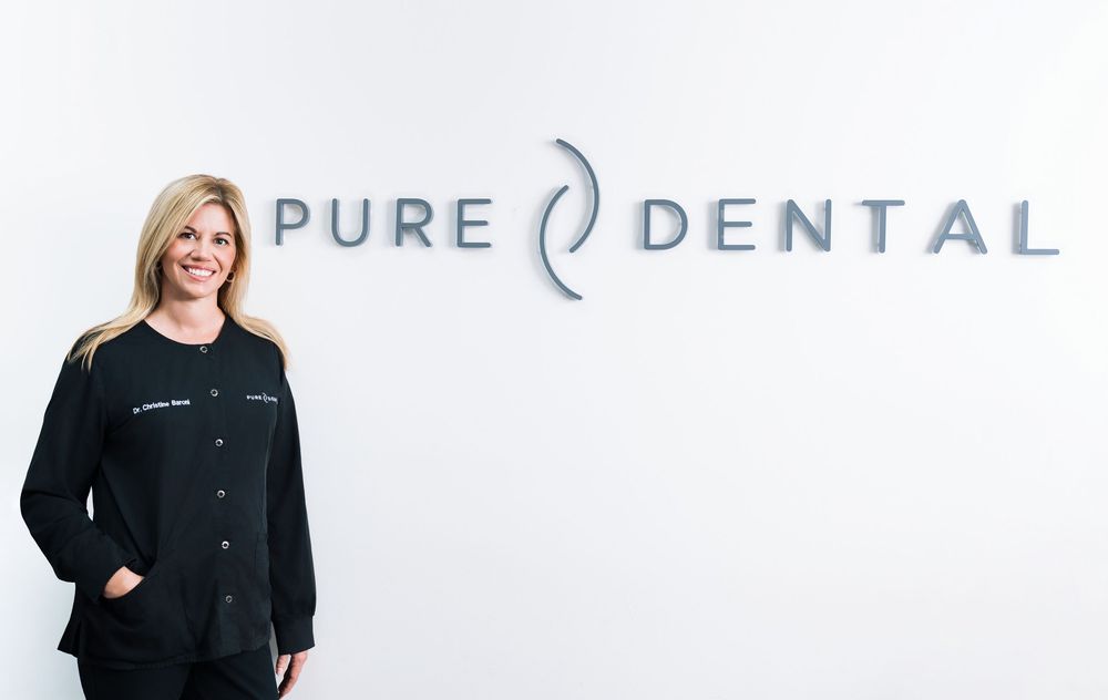 Smiling female dentist in black scrubs standing in front of Pure Dental office sign in modern clinic.