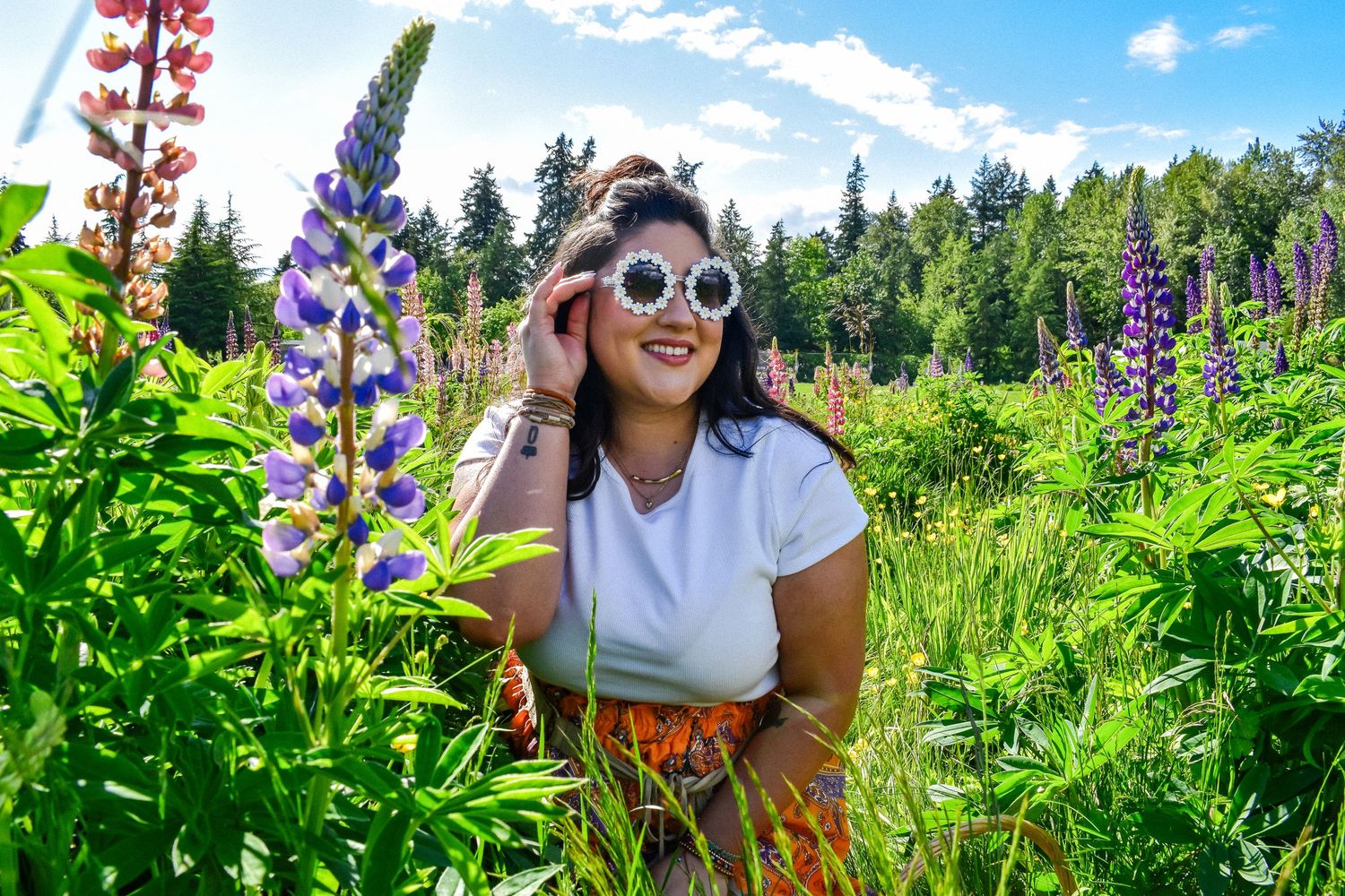 Stylish woman in a flower field wearing sunglasses and a white top, surrounded by tall purple lupines and blue skies. Lupine Session