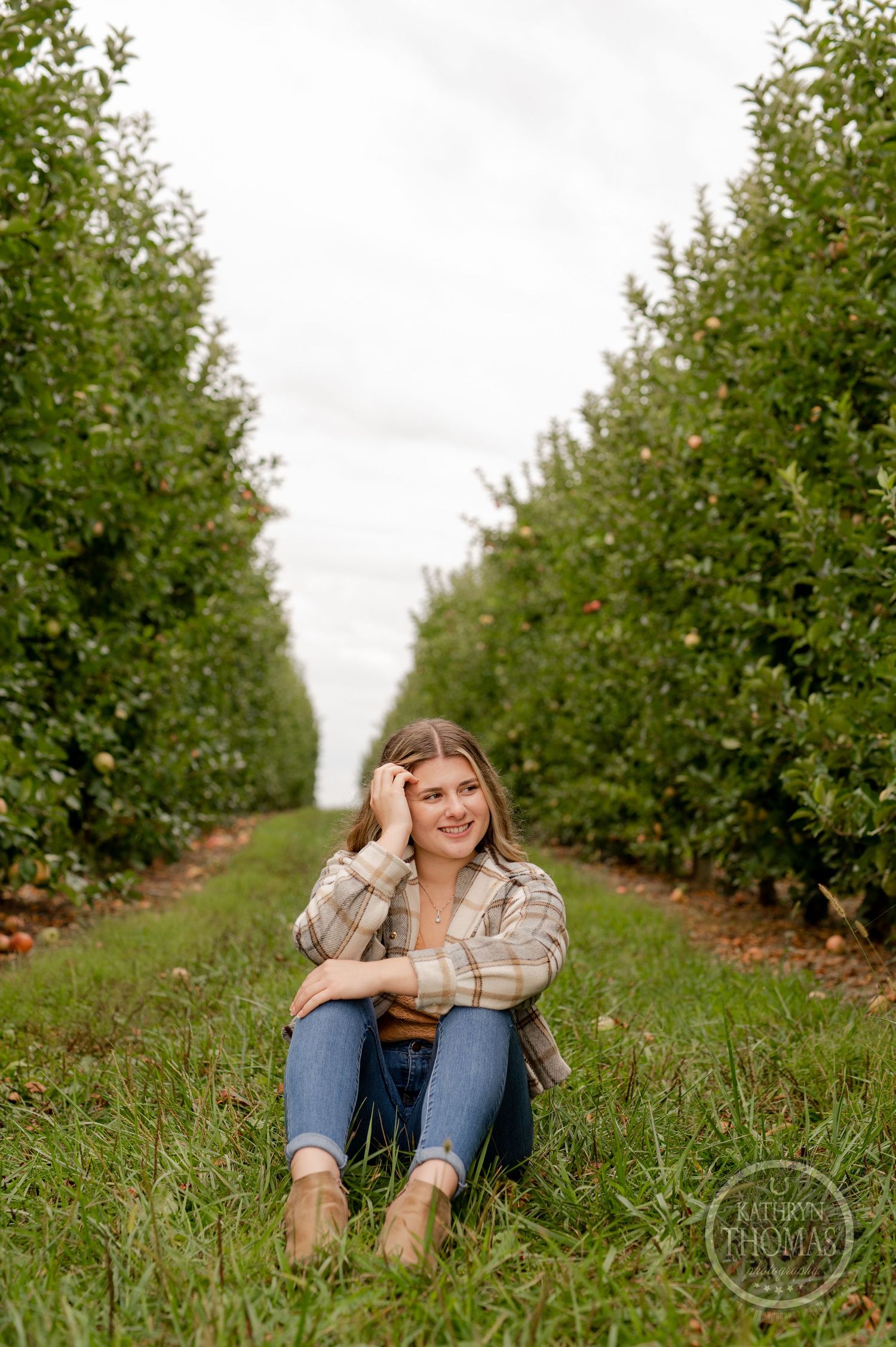 Casual outdoor portrait of someone sitting on grass between rows of apple trees in an orchard.