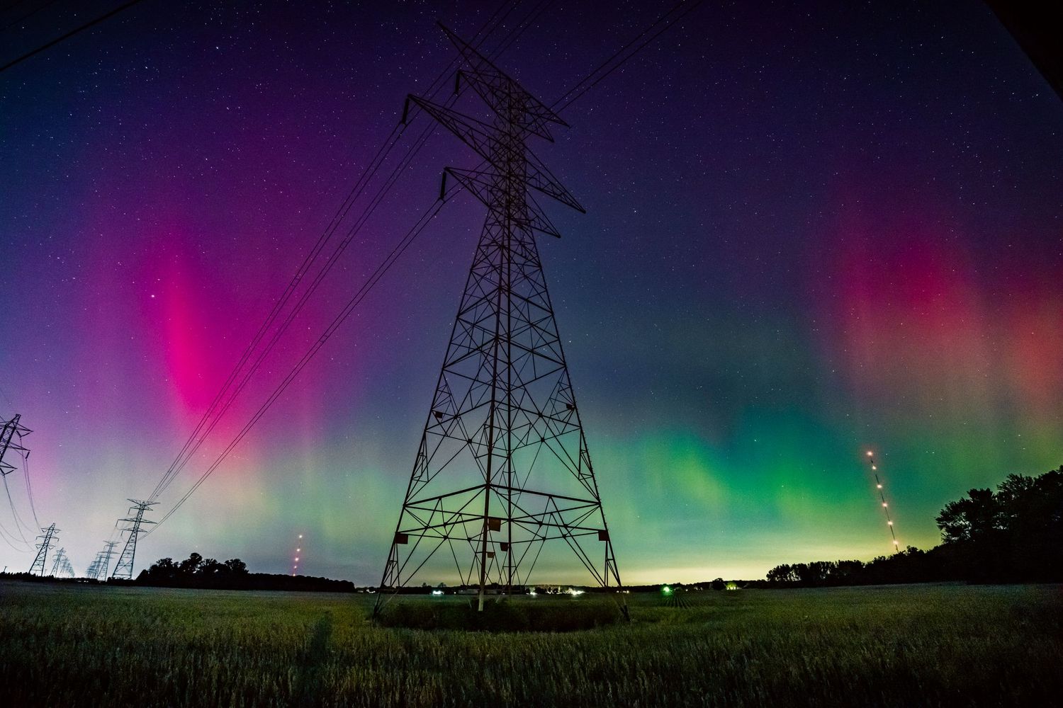 Spectacular aurora borealis lights dancing behind electrical transmission towers in a rural field.