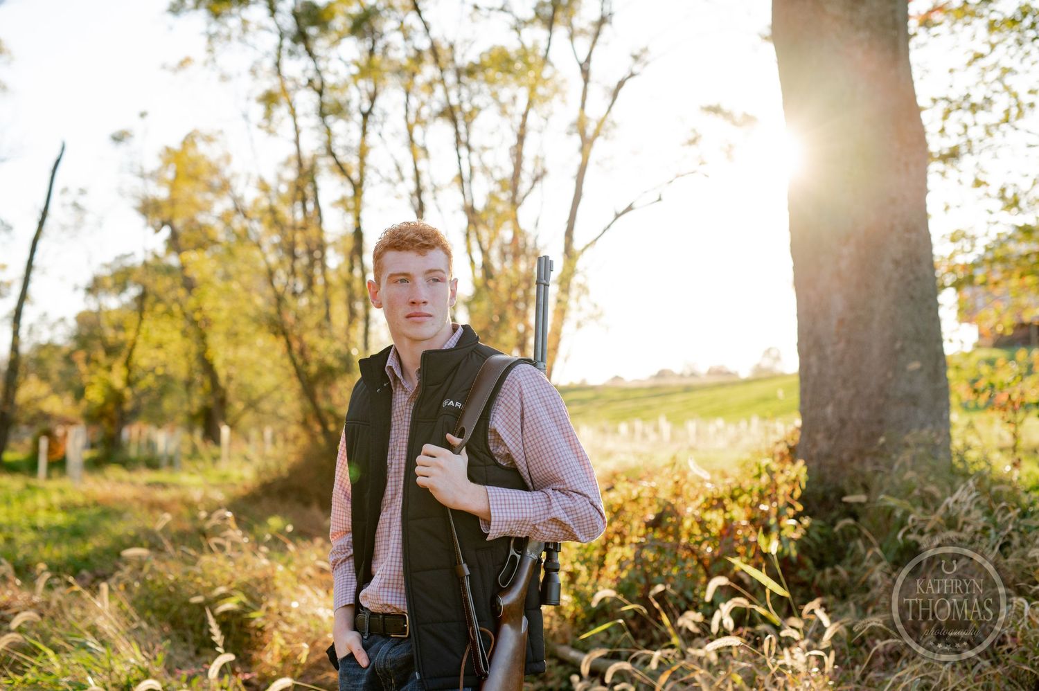 Person standing in sunlit wooded area wearing casual fall attire with sun flare behind trees.