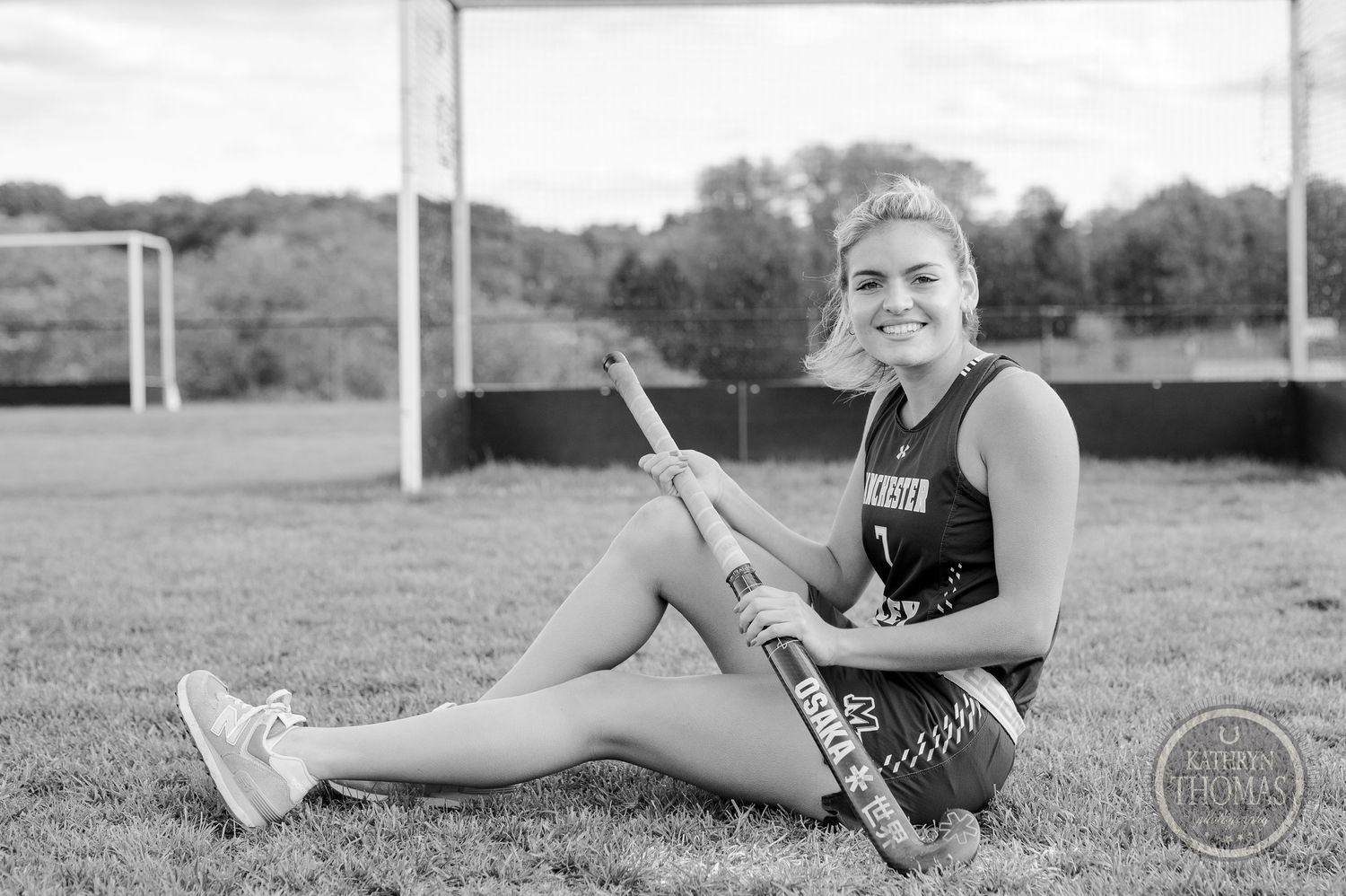 Black and white photo of athlete holding field hockey stick while sitting by goal post.