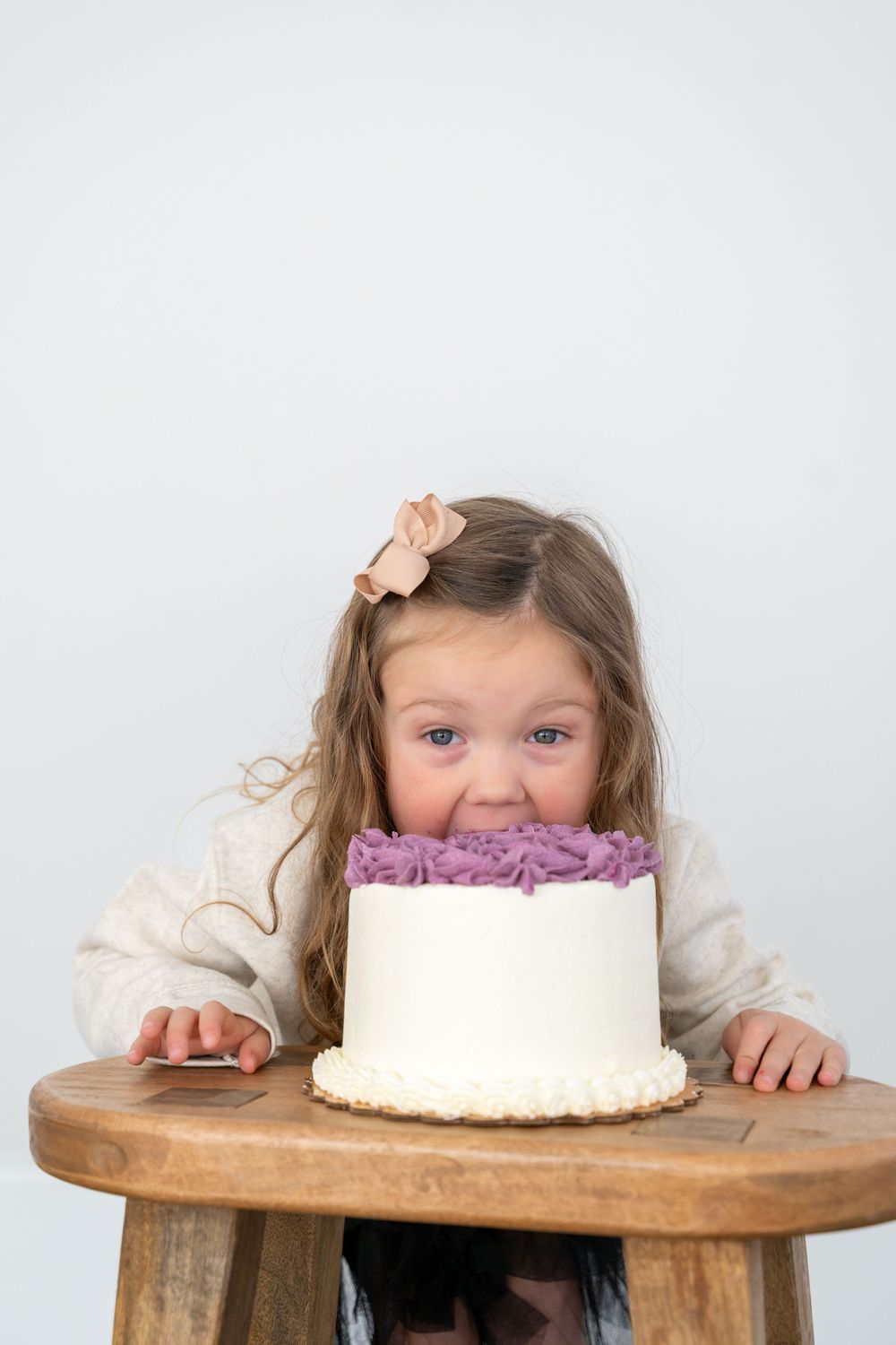 A small child peeks over a wooden table at a white frosted cake with purple decorative details.