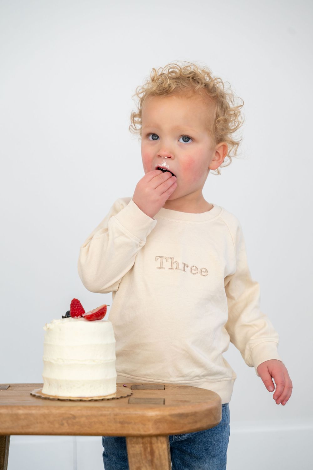 Baby in cream sweater looking curiously at small white birthday cake with red bow on wooden table.