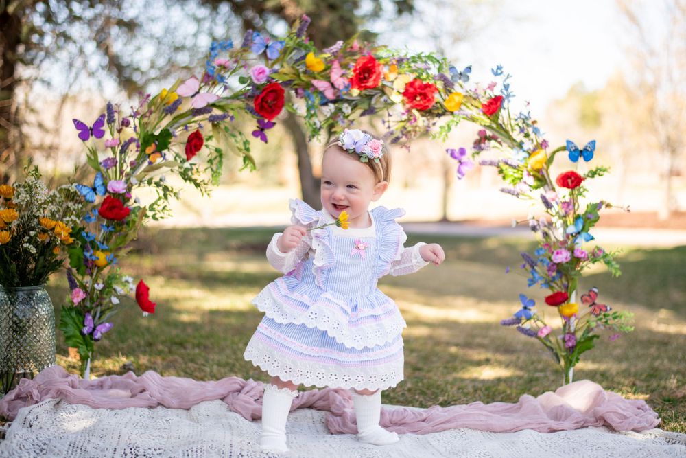 Child in ruffled white dress twirling under floral arch during outdoor photo session. Cake smash