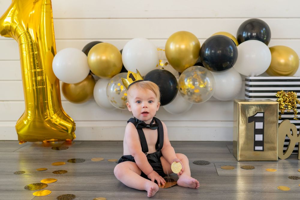 First birthday celebration setup with gold number 1 balloon and black and gold balloon arch against white wall. Cake smash