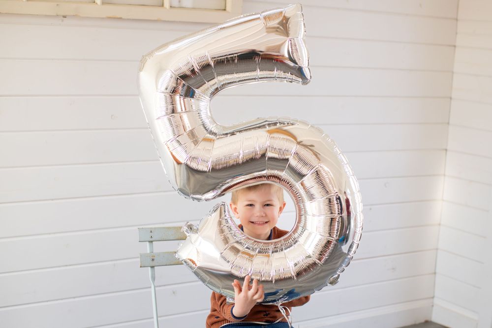 Child holding large silver number 5 balloon against white wall.