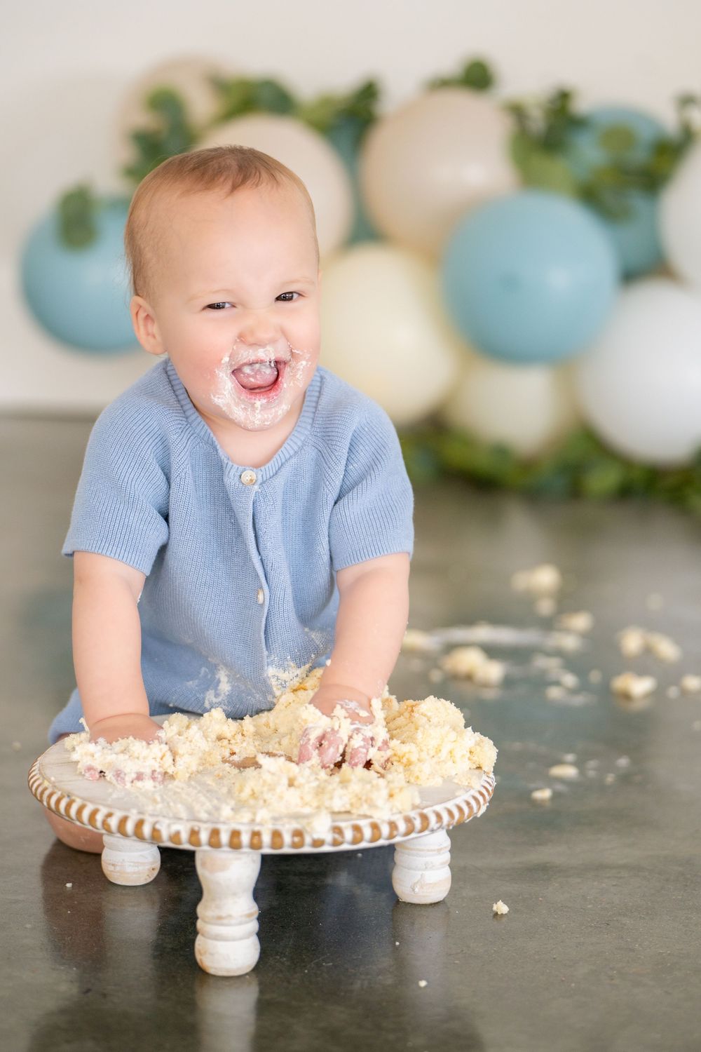A joyful baby in blue sweater plays with cake on a decorative stand with blue and white balloons in background.