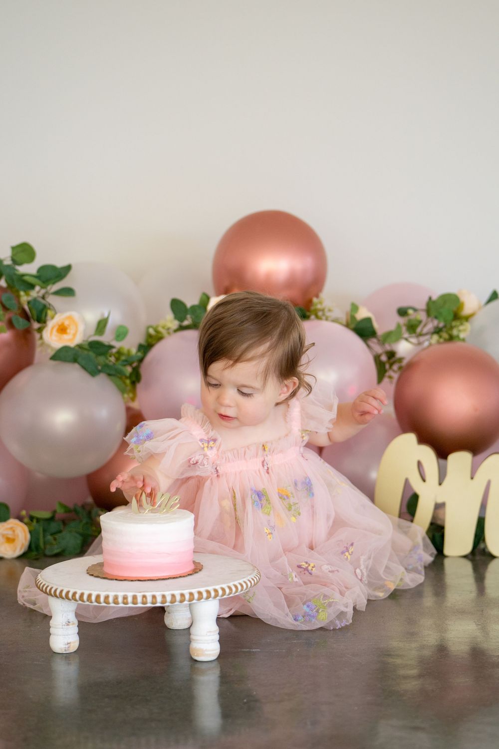 A baby in a pink tulle dress sits on the floor with rose gold and pink balloons and floral decorations in the background.