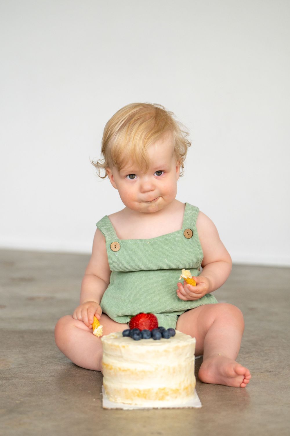 Baby in mint green romper sitting with small cake decorated with berries. Cake smash