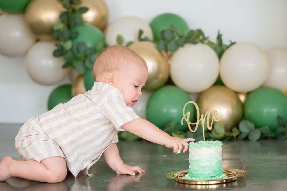 A baby in white clothing reaches for a turquoise birthday cake against a backdrop of green and gold balloons.